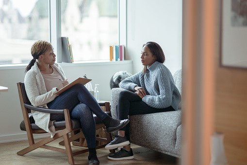 Two people sat in a room in conversation. One is holding a clipboard and pen and talking to the other person.